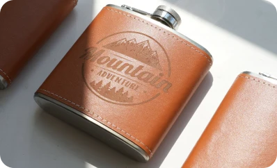 LaserPecker LP2 Plus professional laser engraving on a brown leather hip flask sleeve, featuring a detailed "Mountain Adventure" outdoor-themed logo with deep, clean contrast.