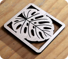 A close-up view of a square wooden coaster featuring a precise laser-cut silhouette of a Monstera leaf, demonstrating the intricate cutting capabilities of the LaserPecker LP2 Plus on wood materials.