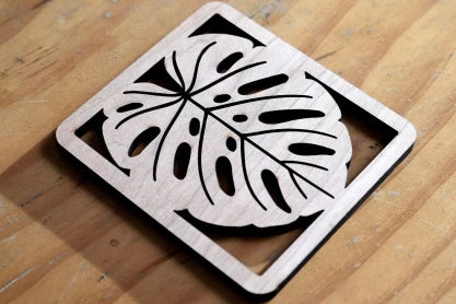 A close-up view of a square wooden coaster featuring a precise laser-cut silhouette of a Monstera leaf, demonstrating the intricate cutting capabilities of the LaserPecker LP2 Plus on wood materials.