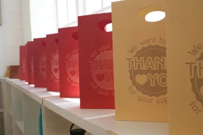 A row of colorful red and tan paper gift bags with laser-engraved "Thank You" logos, showcasing professional-grade branding solutions for boutique owners using LaserPecker LP2 Plus.