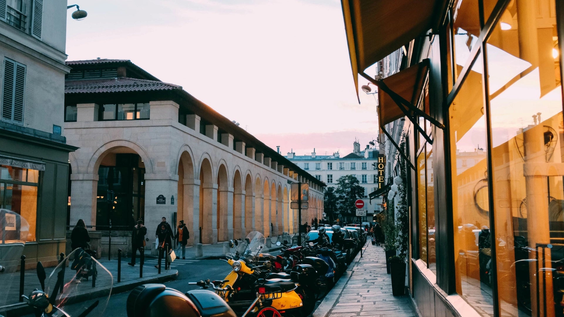 Une scène de rue en France mettant en avant des bâtiments à l’architecture remarquable avec des entrées voûtées rappelant un musée, tandis que des piétons circulent et que des vélos et trottinettes sont soigneusement stationnés le long de la rue.